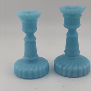 Puede incluir: Dos portavelas de cristal azul claro con acabado translúcido. Cada pieza presenta una base circular ancha con detalles estriados verticales, una columna central con un patrón de espiral y un borde superior acampanado con diseño trenzado.