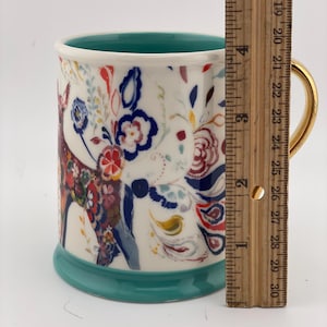 May include: A ceramic mug with a white background, featuring a colorful floral and deer design. The mug has a gold handle, a teal interior, and a teal base. The mug is approximately 4 inches tall.