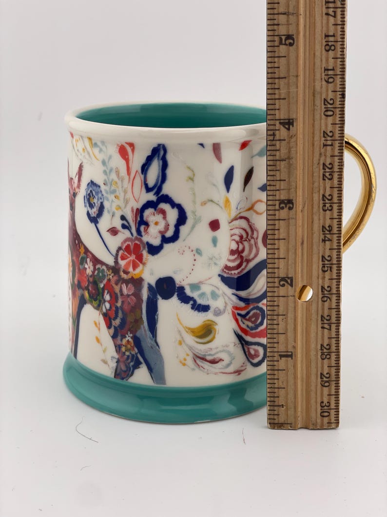 May include: A ceramic mug with a white background, featuring a colorful floral and deer design. The mug has a gold handle, a teal interior, and a teal base. The mug is approximately 4 inches tall.