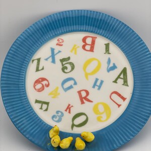May include: A blue ceramic plate with a white center featuring colorful letters and numbers. The plate has a ridged border and is decorated with small yellow chick figurines.