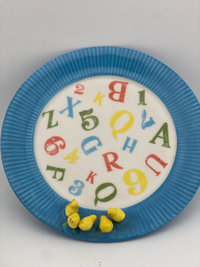 May include: A blue ceramic plate with a white center featuring colorful letters and numbers. The plate has a ridged border and is decorated with small yellow chick figurines.
