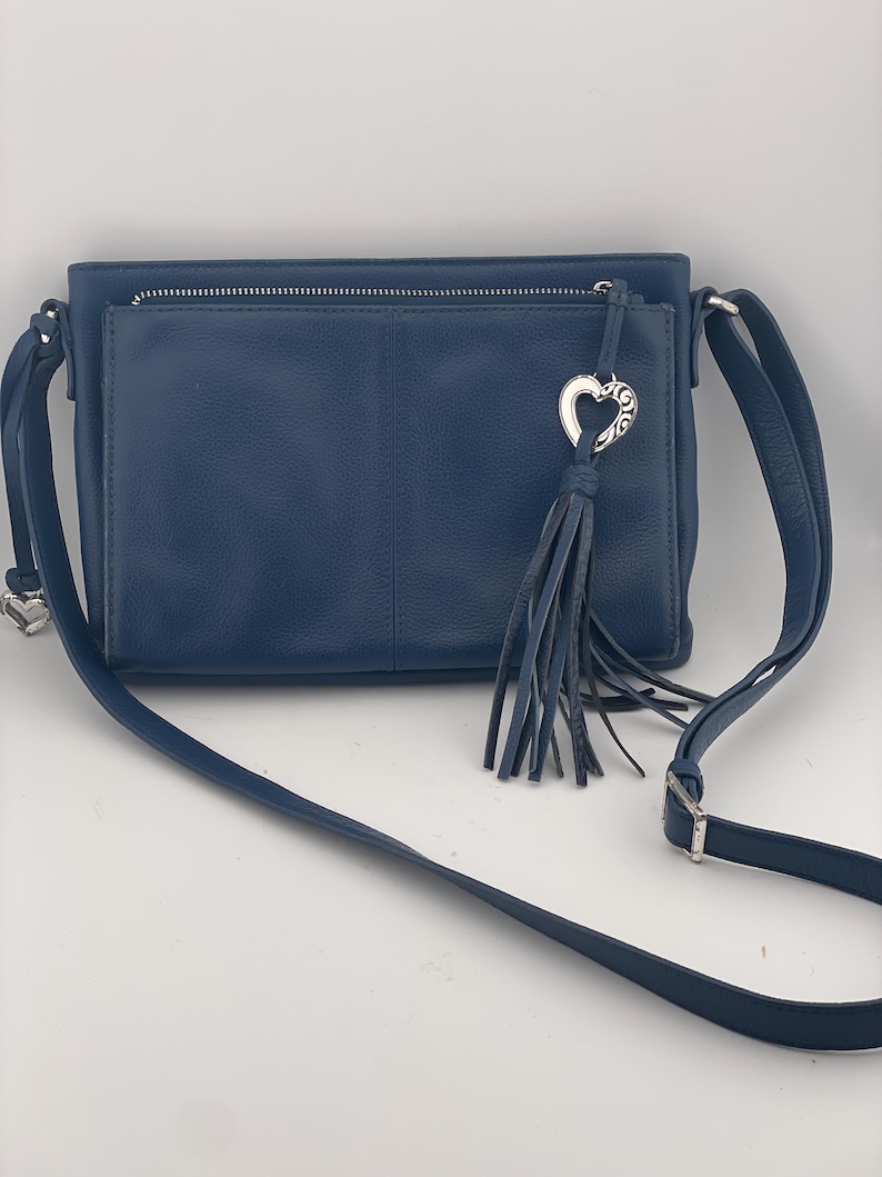 May include: A navy blue crossbody bag with a front pocket and a silver zipper. The bag features a silver heart-shaped charm with a tassel. The adjustable strap is also navy blue.