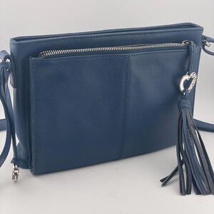 May include: A navy blue crossbody bag with a front pocket and a zippered top. The bag features a silver zipper and hardware, a tassel, and a heart-shaped charm. The bag has a textured appearance and a long strap.