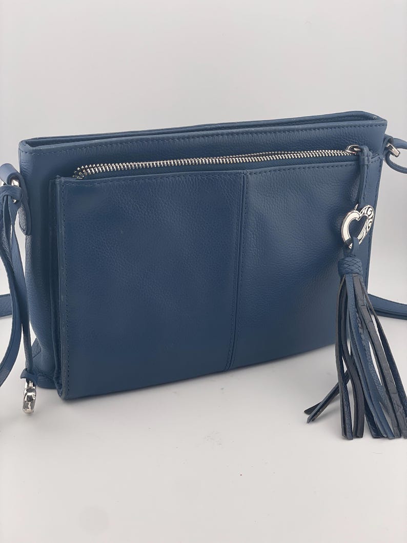 May include: A navy blue crossbody bag with a front pocket and a zippered top. The bag features a silver zipper and hardware, a tassel, and a heart-shaped charm. The bag has a textured appearance and a long strap.