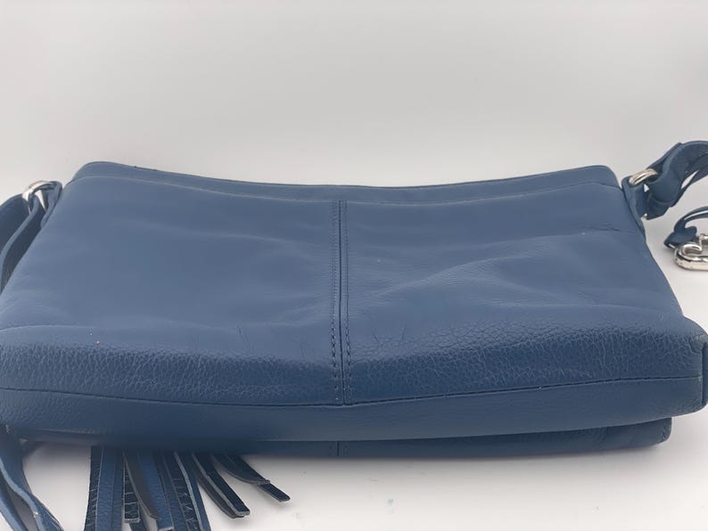 May include: A navy blue leather crossbody bag with a long adjustable strap and silver-tone hardware. The bag features a front flap with fringe detailing and a stitched center seam. The bag is designed for everyday use.