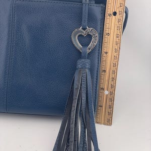 May include: A blue leather handbag with a decorative heart-shaped charm and a tassel. The bag has a zipper closure and a textured surface. The ruler shows the bag is approximately 12 inches long.