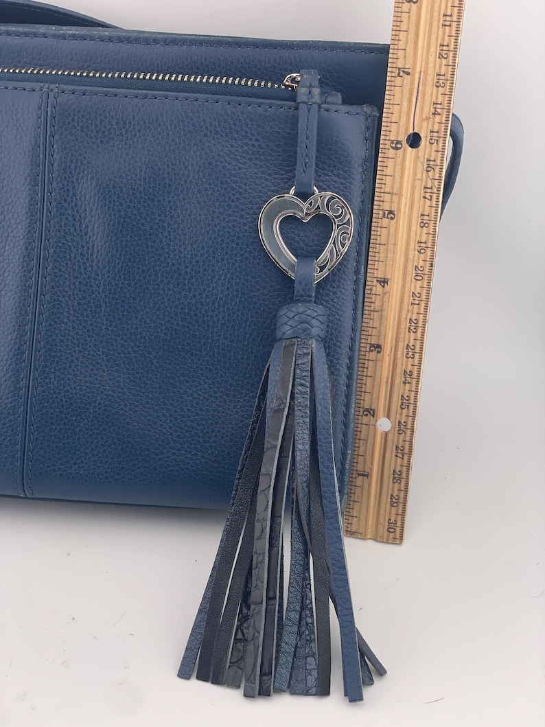May include: A blue leather handbag with a decorative heart-shaped charm and a tassel. The bag has a zipper closure and a textured surface. The ruler shows the bag is approximately 12 inches long.