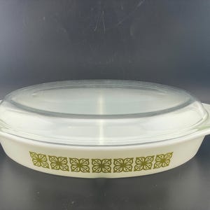 May include: An oval white glass casserole dish with integrated side handles and a clear glass lid. The exterior of the dish features a repeating pattern of stylized green floral motifs arranged in a horizontal band around the center.