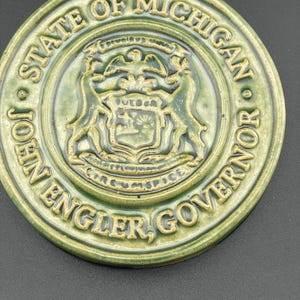 Könnte beinhalten: Ein grüner Keramikuntersetzer mit dem Staatssiegel von Michigan und dem Text "STATE OF MICHIGAN JOHN ENGLER, GOVERNOR".