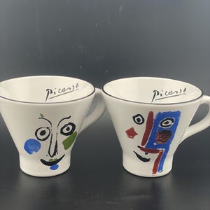 Op de afbeelding: Twee witte keramische mokken met abstracte gezichten in blauw, groen, rood en zwart. Elke mok heeft een uitlopende rand, een handvat en het woord Picasso in zwarte letters aan de binnenrand. Het ontwerp is minimalistisch en kleurrijk.