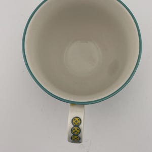 May include: A white ceramic coffee mug with a teal rim and handle. The handle is decorated with three yellow and blue circular designs. The mug is empty and sits on a white surface.