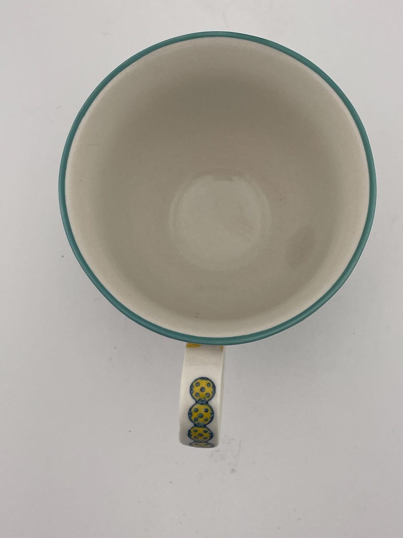 May include: A white ceramic coffee mug with a teal rim and handle. The handle is decorated with three yellow and blue circular designs. The mug is empty and sits on a white surface.