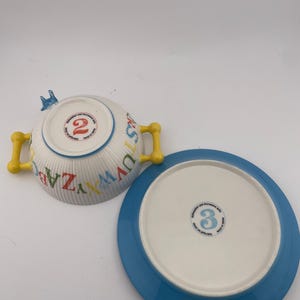 May include: A white ceramic bowl with yellow handles and a matching blue-rimmed plate. The bowl is decorated with colorful letters and the number "2". The plate has the number "3" on the bottom. The set is designed for children.