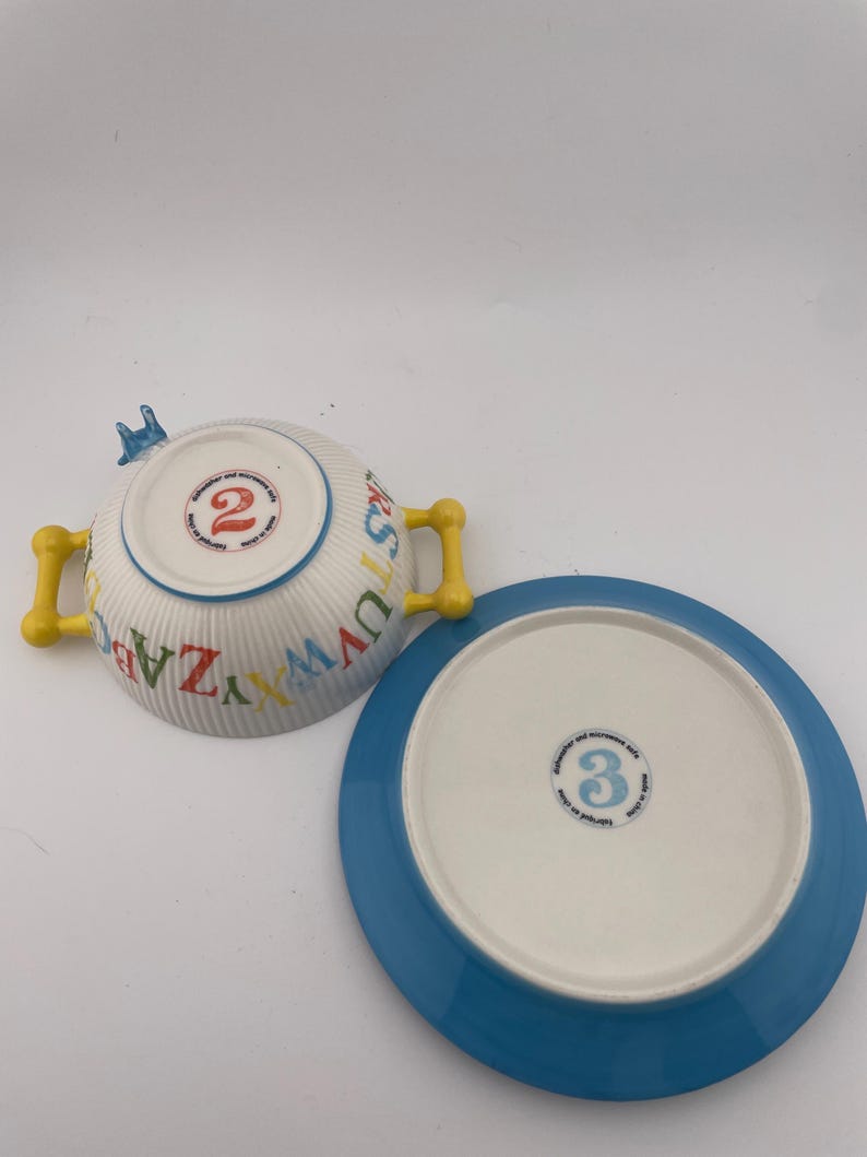 May include: A white ceramic bowl with yellow handles and a matching blue-rimmed plate. The bowl is decorated with colorful letters and the number "2". The plate has the number "3" on the bottom. The set is designed for children.