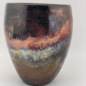 May include: A handcrafted ceramic vase with a wide, rounded body and a dark, glossy finish. The vase features a blend of colors, including copper, brown, and hints of blue and yellow, creating a unique, textured appearance. The vase is suitable for home decor.