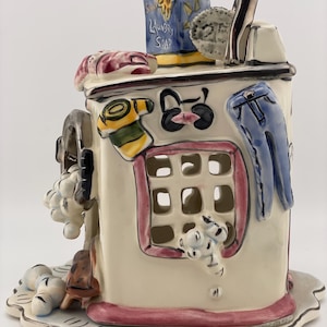 May include: A quirky ceramic laundry room ornament. The off-white structure features a red-trimmed window, a blue denim-like element, and a laundry soap container. Decorative suds and other laundry-themed details adorn the piece, creating a playful aesthetic.
