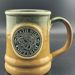 May include: A ceramic coffee mug with a green and brown glaze. The mug has a large, round logo on the front that says "Death Wish Coffee Co." in gold lettering with a black outline. The logo features a stylized image of a dragon.