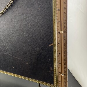 May include: A vintage black framed picture with a gold trim and chain. The frame has small legs and a ruler is placed next to it. The ruler measures up to 30 inches.