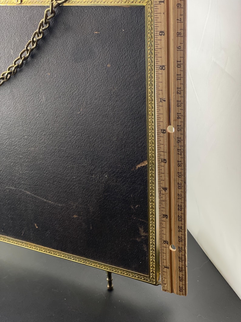 May include: A vintage black framed picture with a gold trim and chain. The frame has small legs and a ruler is placed next to it. The ruler measures up to 30 inches.