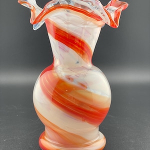 Może przedstawiać: Biały i pomarańczowy szklany wazon o spiralnym wzorze. Wazon ma szeroki, falbankowy otwór i okrągłe ciało.
