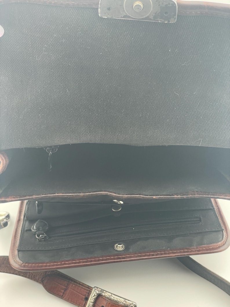 May include: Open view of a brown leather crossbody bag with a textured strap and silver buckle. The bag's interior is black, with a zippered pocket and a magnetic closure on the flap.