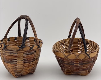 Puede incluir: Dos pequeñas cestas tejidas a mano con asas arqueadas. Las cestas están hechas de material tejido de color natural con detalles en marrón oscuro. Las asas también están tejidas y unidas al borde de la cesta.