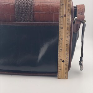 May include: A black and brown leather handbag with a woven strap and a textured, crocodile-like pattern. The bag is approximately 25 cm in length.