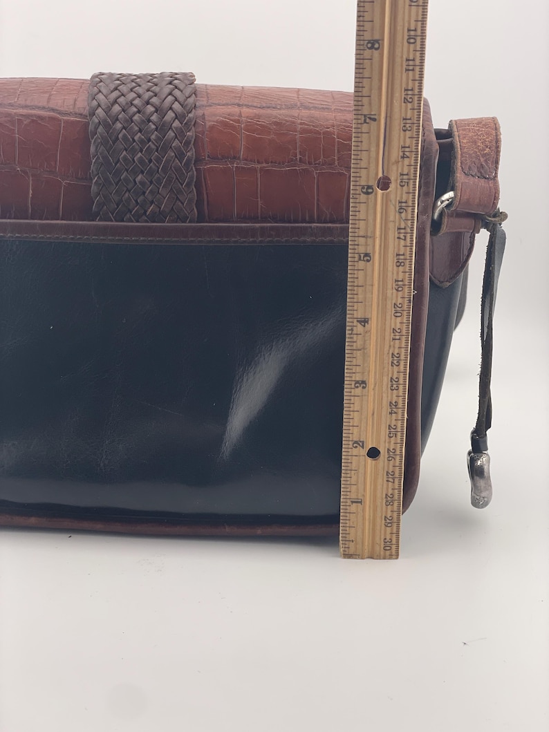 May include: A black and brown leather handbag with a woven strap and a textured, crocodile-like pattern. The bag is approximately 25 cm in length.
