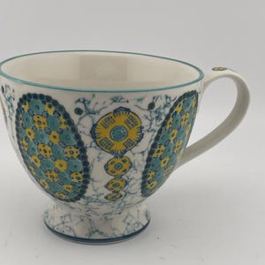 May include: White ceramic teacup with a large handle and a decorative pattern in teal and yellow. The pattern features floral and geometric shapes. The rim and base of the cup are trimmed in teal.