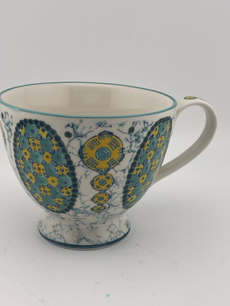 May include: White ceramic teacup with a large handle and a decorative pattern in teal and yellow. The pattern features floral and geometric shapes. The rim and base of the cup are trimmed in teal.
