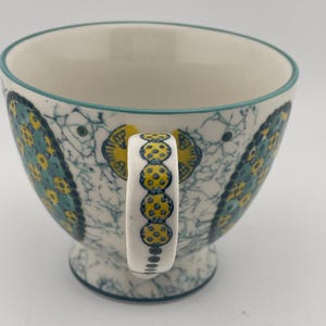 May include: A ceramic teacup with a white interior and a teal rim. The exterior features a blue and yellow floral pattern on a white background. The handle is white with yellow polka dots. The cup sits on a small pedestal base.