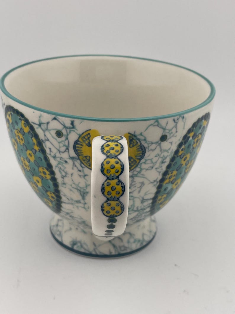 May include: A ceramic teacup with a white interior and a teal rim. The exterior features a blue and yellow floral pattern on a white background. The handle is white with yellow polka dots. The cup sits on a small pedestal base.