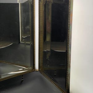 May include: A vintage vanity mirror with a brass frame and mirrored panels. The mirror has a decorative design around the edges and stands on small, ornate legs. The mirror is open, revealing the reflective surfaces.