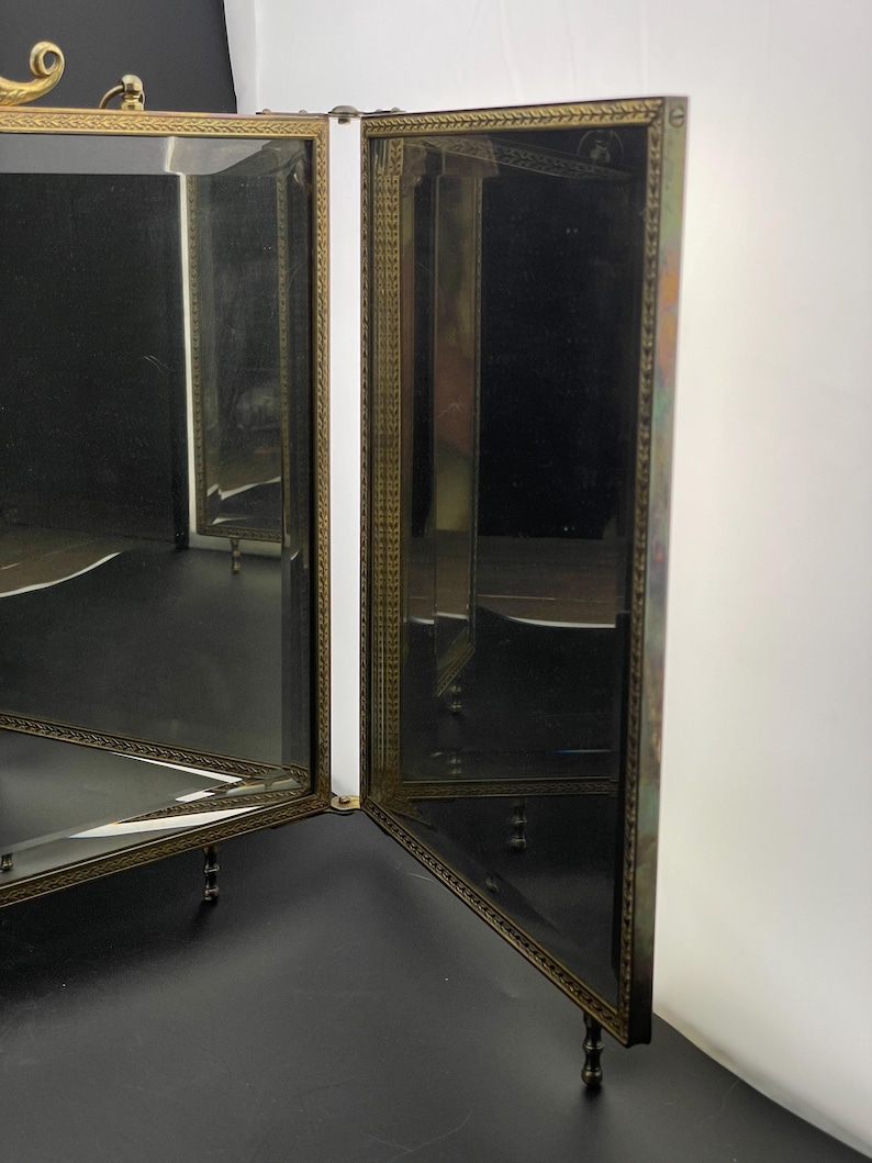 May include: A vintage vanity mirror with a brass frame and mirrored panels. The mirror has a decorative design around the edges and stands on small, ornate legs. The mirror is open, revealing the reflective surfaces.