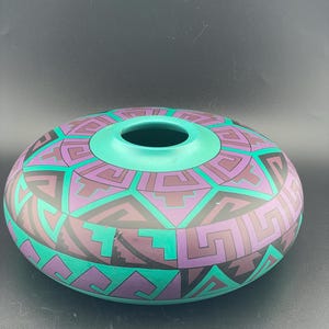May include: A ceramic, round vase with a wide opening and a flat base. The vase is painted with a geometric pattern in teal, purple, and black. The opening and the base are teal.