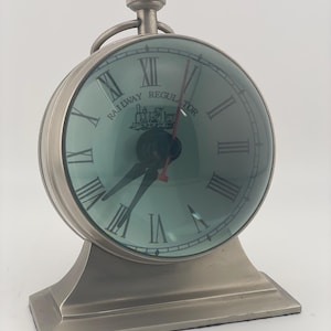 May include: A silver-toned metal table clock with a round face and a wide, rectangular base. The clock features a light blue dial with black Roman numerals, a central steam locomotive graphic, and the text RAILWAY REGULATOR. It has black hour and minute hands and a thin red second hand.