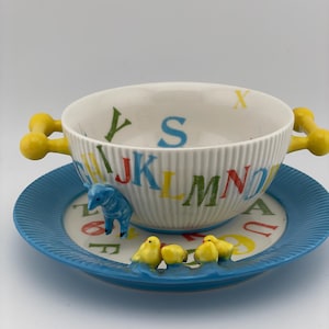 May include: A white ceramic bowl and saucer set with a playful alphabet design. The bowl has yellow handles and is decorated with colorful letters. The blue saucer features a blue elephant and yellow chicks.