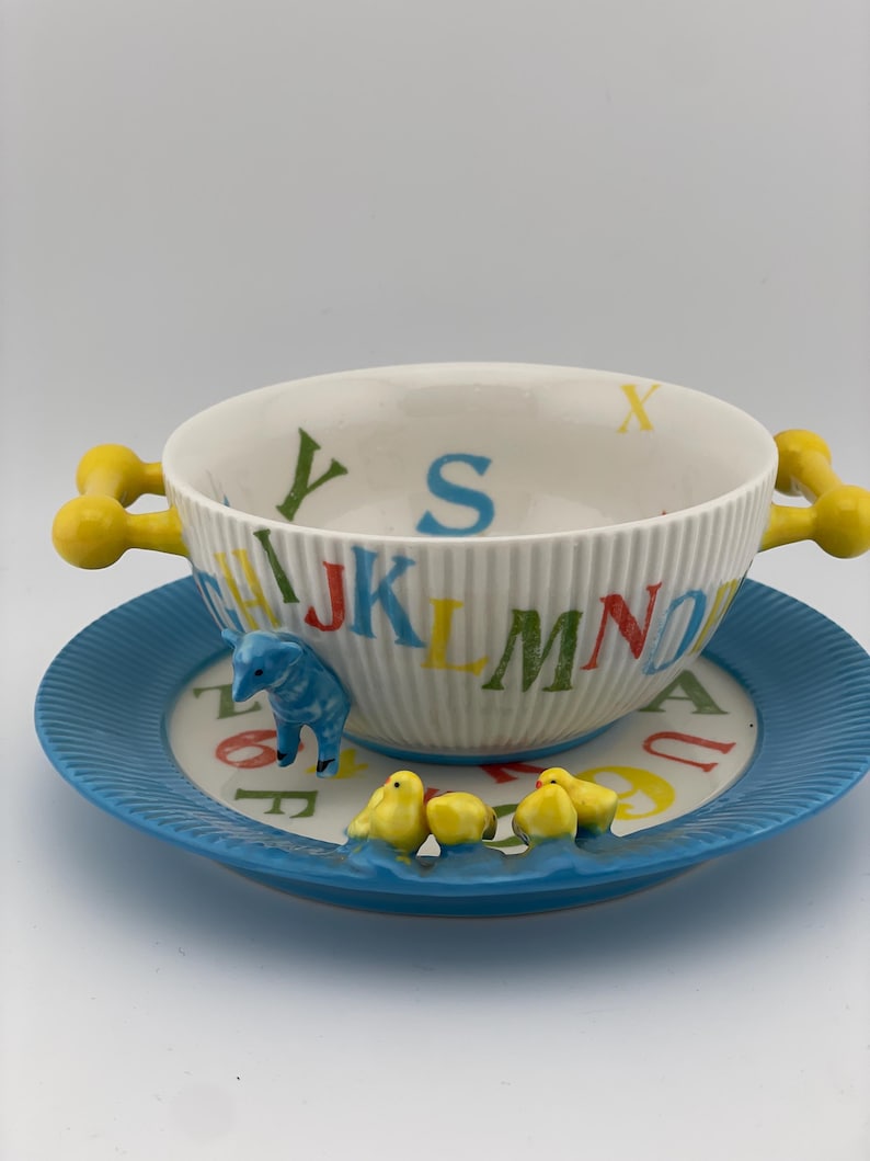 May include: A white ceramic bowl and saucer set with a playful alphabet design. The bowl has yellow handles and is decorated with colorful letters. The blue saucer features a blue elephant and yellow chicks.