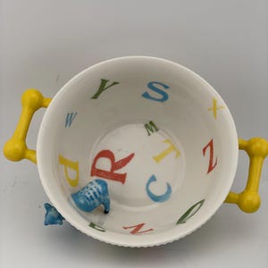 May include: A white ceramic bowl with yellow handles. The interior features colorful letters of the alphabet and a small blue animal figurine. The bowl is designed for serving and decorative purposes.