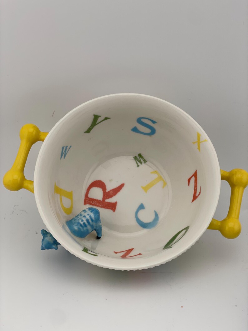 May include: A white ceramic bowl with yellow handles. The interior features colorful letters of the alphabet and a small blue animal figurine. The bowl is designed for serving and decorative purposes.