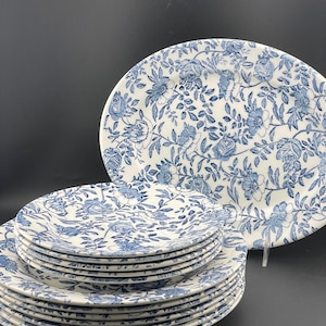 May include: A collection of white ceramic plates and an oval serving platter, adorned with a blue floral design. The plates are stacked, and the platter is oval. The floral pattern includes roses and leaves, offering a classic, elegant aesthetic. The background is black.