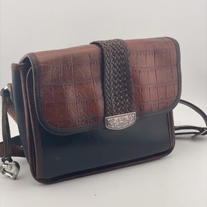 May include: A brown and black crossbody bag with a textured, crocodile-like pattern. The bag features a braided strap closure and a silver-toned decorative clasp. The strap has a silver heart-shaped charm.