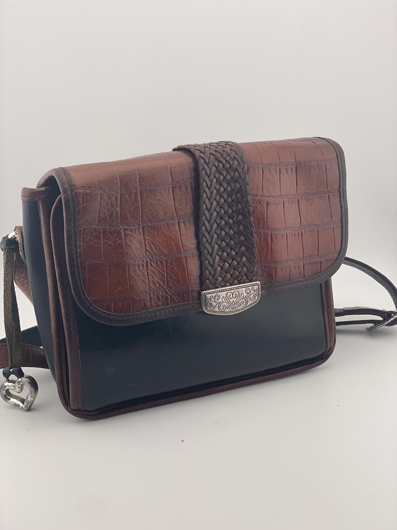 May include: A brown and black crossbody bag with a textured, crocodile-like pattern. The bag features a braided strap closure and a silver-toned decorative clasp. The strap has a silver heart-shaped charm.