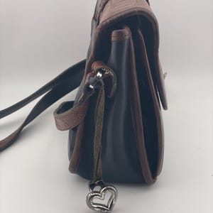May include: A black and brown leather crossbody bag with a decorative heart charm. The bag features a textured flap and a braided detail on the top. The strap is black, and the trim is brown.