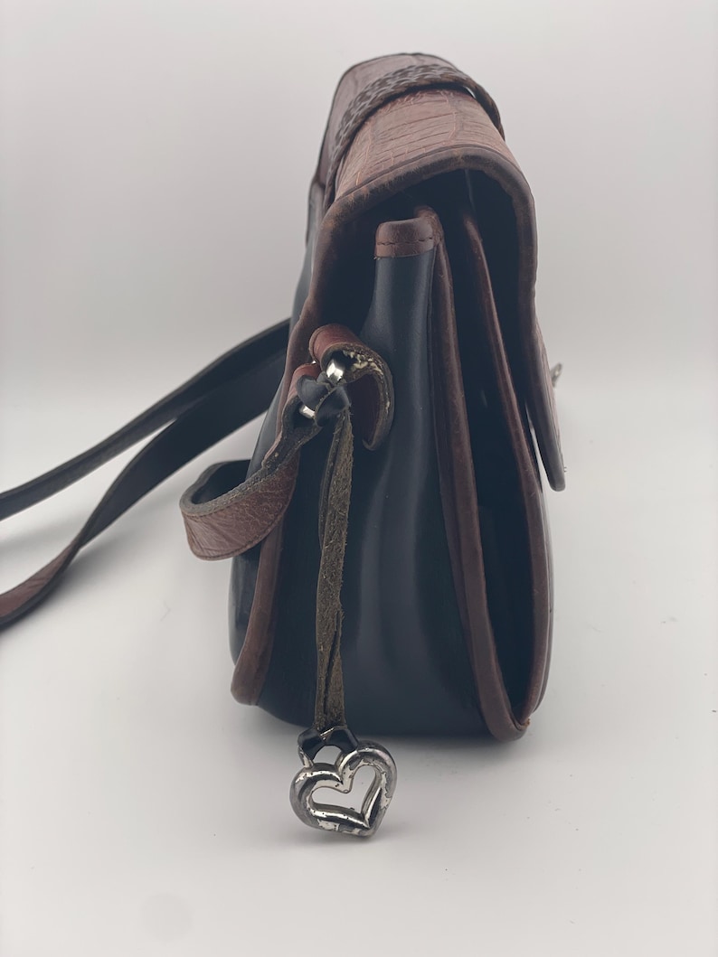 May include: A black and brown leather crossbody bag with a decorative heart charm. The bag features a textured flap and a braided detail on the top. The strap is black, and the trim is brown.