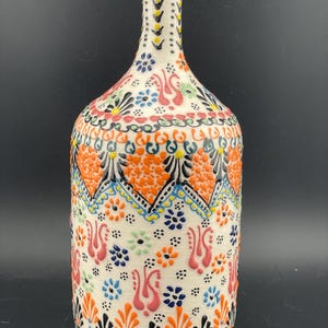 Może przedstawiać: Dekoracyjna ceramiczna butelka z wąską szyjką i zaokrąglonym korpusem. Butelka zdobiona jest kolorowym wzorem kwiatowym w kolorze pomarańczowym, niebieskim, różowym i czarnym na białym tle. Wzór zawiera stylizowane kwiaty i kształty geometryczne.