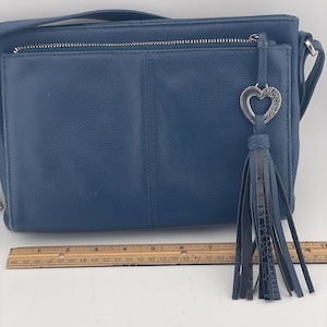 May include: A blue leather crossbody bag with a silver zipper and a decorative heart-shaped charm. The bag features a long, dark blue tassel and a shoulder strap. The bag is approximately 10 inches wide.