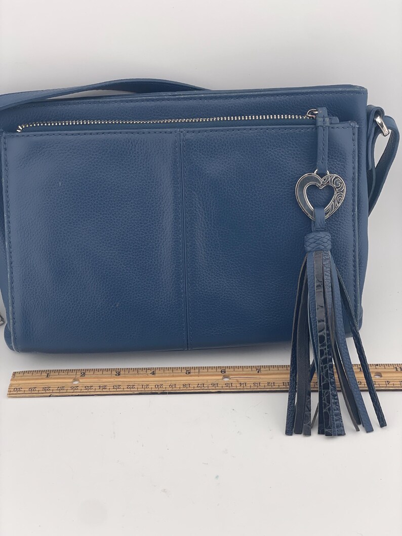 May include: A blue leather crossbody bag with a silver zipper and a decorative heart-shaped charm. The bag features a long, dark blue tassel and a shoulder strap. The bag is approximately 10 inches wide.
