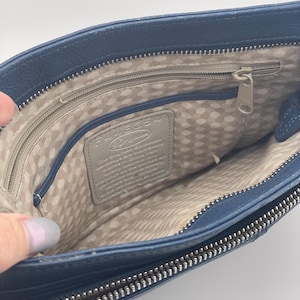 May include: Open view of a navy blue leather handbag with a silver zipper. The interior features a light beige polka dot lining and a gray leather tag. The bag is held open, showcasing the inside.