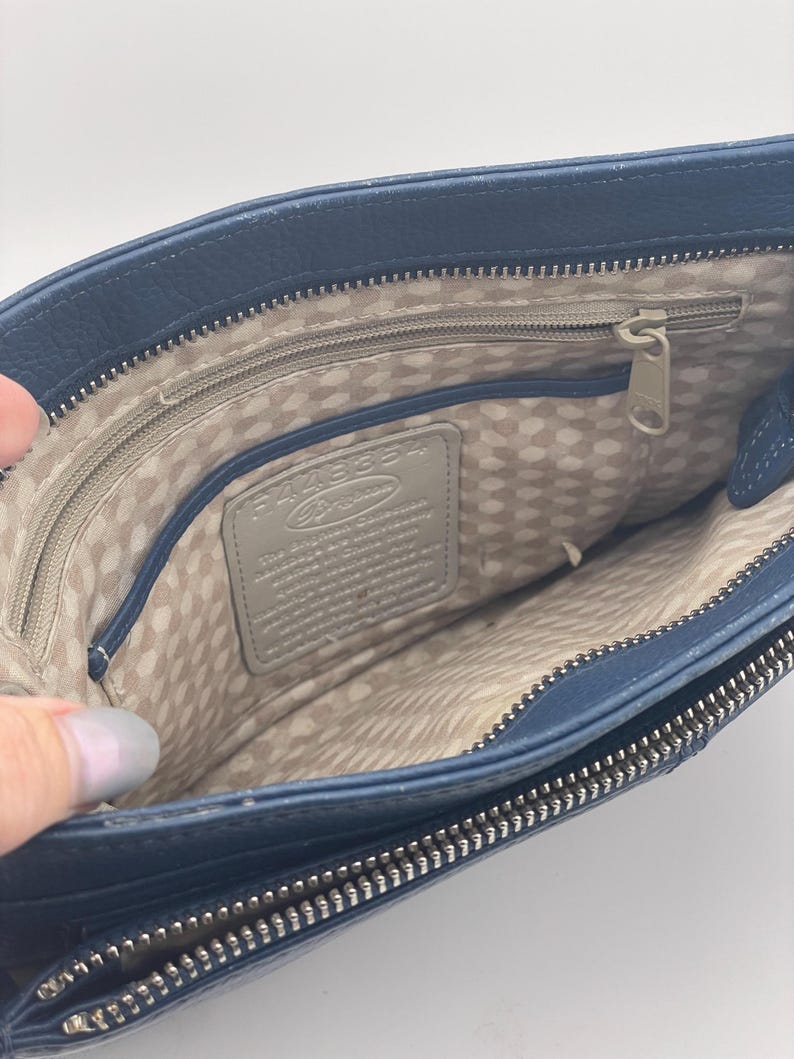 May include: Open view of a navy blue leather handbag with a silver zipper. The interior features a light beige polka dot lining and a gray leather tag. The bag is held open, showcasing the inside.